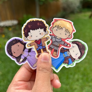May include: Four cartoon stickers featuring characters in various outfits. One sticker shows a character in a purple dress, another in a blue outfit, and two others in brown and blue outfits. The stickers have a white border and are held in a hand.