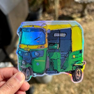 May include: A colorful sticker of an auto rickshaw, featuring green, yellow, and blue hues. The sticker has the text "KHOWP 6845K" and "HYDERABADBADIE". The image is held by a hand.