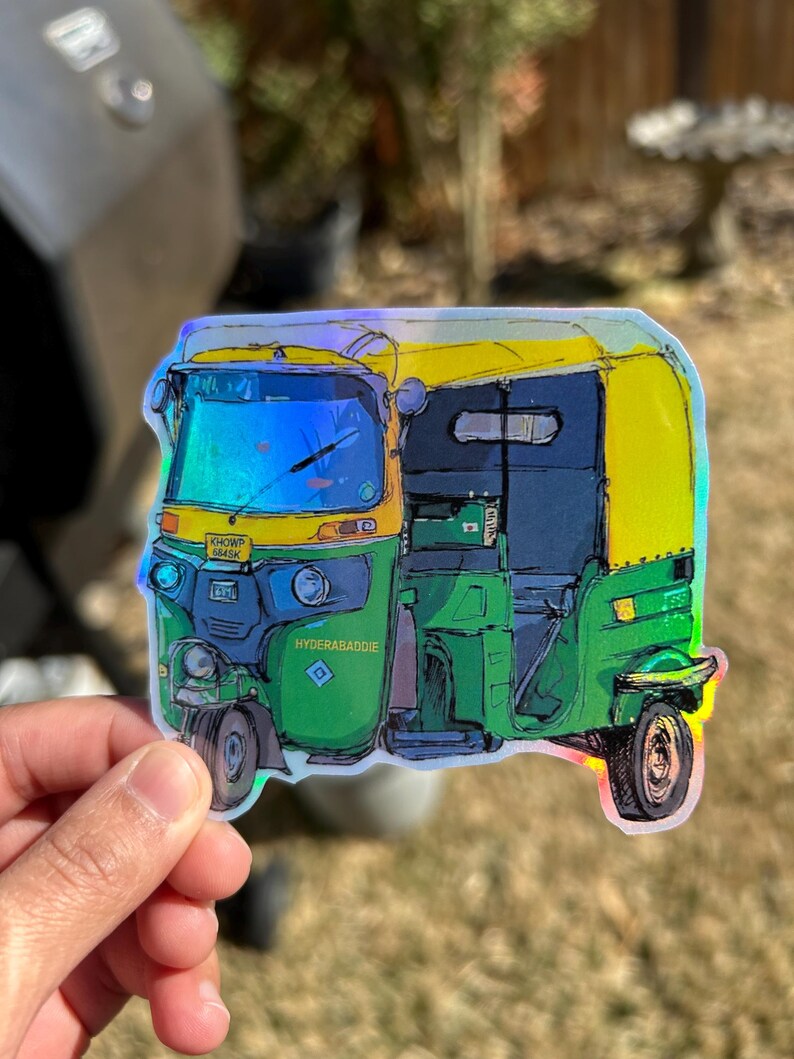 May include: A colorful sticker of an auto rickshaw, featuring green, yellow, and blue hues. The sticker has the text "KHOWP 6845K" and "HYDERABADBADIE". The image is held by a hand.