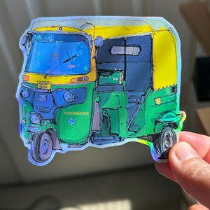 May include: A colorful sticker featuring an auto-rickshaw, a three-wheeled vehicle. The sticker has a holographic effect and features green, yellow, and blue colors. The text "HYDERABADDIE" is visible on the side of the vehicle.