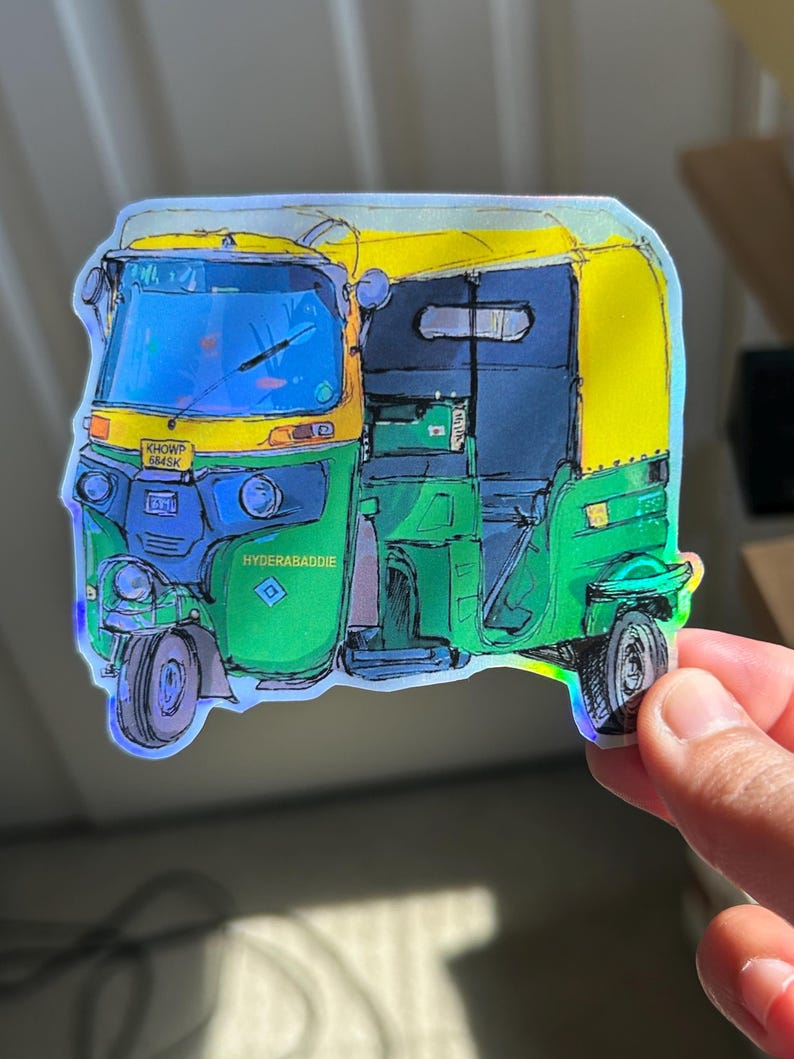 May include: A colorful sticker featuring an auto-rickshaw, a three-wheeled vehicle. The sticker has a holographic effect and features green, yellow, and blue colors. The text "HYDERABADDIE" is visible on the side of the vehicle.