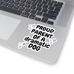 Puede incluir: Un portátil blanco con una pegatina negra que dice "Proud Parent of a Dramatic Dog" con pequeñas estrellas alrededor del texto.