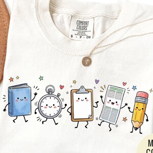 May include: An ivory t-shirt featuring a graphic print of five anthropomorphic school supplies including a blue book, a silver stopwatch, a brown clipboard, a white test paper, and a yellow pencil, all with smiling faces and limbs.