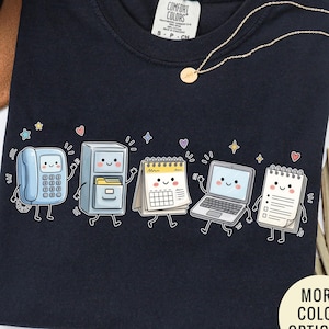 May include: A black short-sleeved t-shirt featuring a graphic print of five anthropomorphic office items including a telephone, filing cabinet, calendar, laptop, and notepad, each with a smiling face, surrounded by small colorful stars and hearts.
