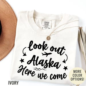 May include: Ivory-colored t-shirt with the text "look out Alaska Here we come" in black script. The design includes a plane, stars, and waves. A gold necklace and a brown hat are also visible.