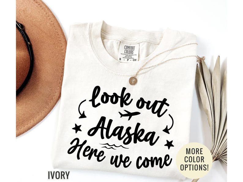 May include: Ivory-colored t-shirt with the text "look out Alaska Here we come" in black script. The design includes a plane, stars, and waves. A gold necklace and a brown hat are also visible.