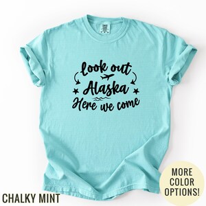 May include: A light blue t-shirt with the text "look out Alaska Here we come" in black. The design includes a plane, stars, and waves. The shirt is a chalky mint color.