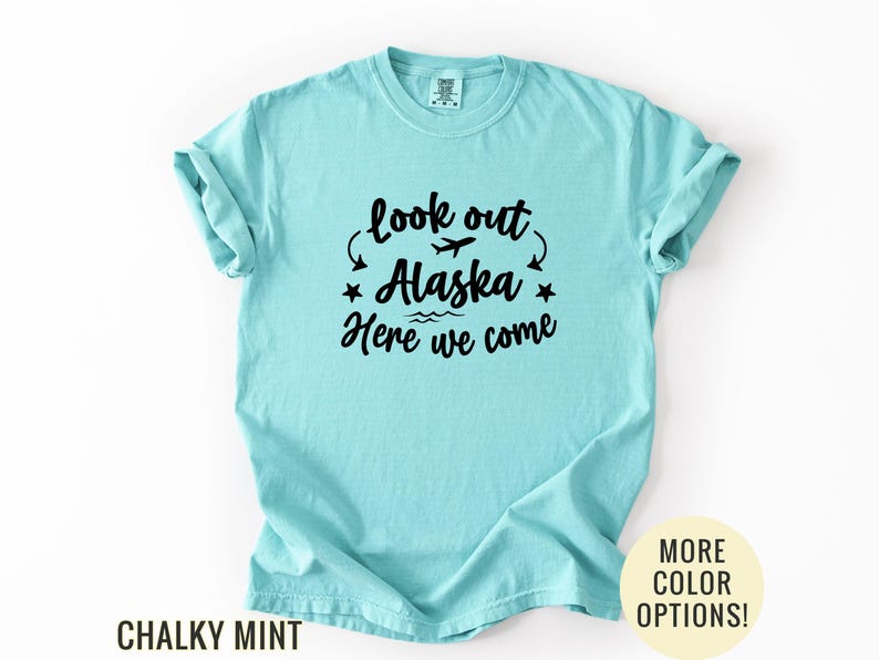 May include: A light blue t-shirt with the text "look out Alaska Here we come" in black. The design includes a plane, stars, and waves. The shirt is a chalky mint color.