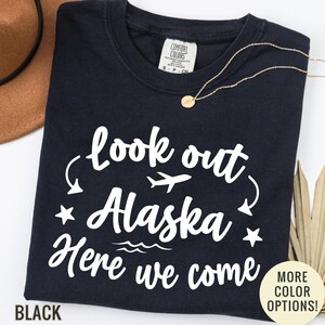 May include: Black t-shirt with the text "look out Alaska Here we come" in white script. The design includes a plane, stars, and waves. A gold necklace and a brown hat are also visible.