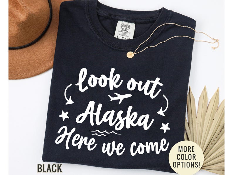 May include: Black t-shirt with the text "look out Alaska Here we come" in white script. The design includes a plane, stars, and waves. A gold necklace and a brown hat are also visible.