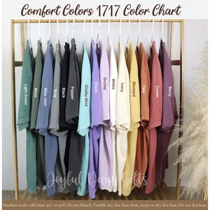 May include: A color chart displays Comfort Colors 1717 t-shirts in various hues. The shirts are arranged on a wooden rack, showcasing colors like Light Green, Moss, Blue Jean, Black, and Berry. The text "Comfort Colors 1717 Color Chart" is at the top.