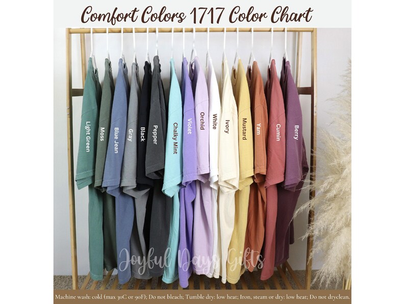 May include: A color chart displays Comfort Colors 1717 t-shirts in various hues. The shirts are arranged on a wooden rack, showcasing colors like Light Green, Moss, Blue Jean, Black, and Berry. The text "Comfort Colors 1717 Color Chart" is at the top.