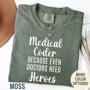 May include: A moss green short-sleeve t-shirt with white text that reads Medical Coder BECAUSE EVEN DOCTORS NEED Heroes. The shirt features a crew neckline and a Comfort Colors brand label on the inside collar, indicating a size small.