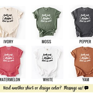 May include: A collection of six t-shirts in various colors: ivory, moss, pepper, watermelon, white, and yam. Each shirt features the text "look out Alaska, Here we come" in a decorative font. The bottom of the image has the text "Need another shirt or design color? Message us!"