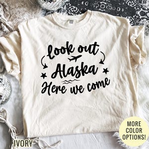 May include: Ivory-colored t-shirt with the text "look out Alaska Here we come" in black. The design includes an airplane, stars, and waves. The shirt has short sleeves and a crew neck.