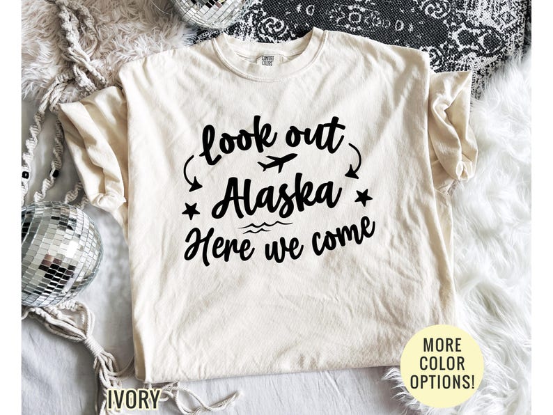 May include: Ivory-colored t-shirt with the text "look out Alaska Here we come" in black. The design includes an airplane, stars, and waves. The shirt has short sleeves and a crew neck.