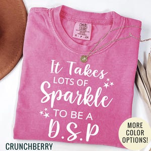 May include: A pink short-sleeved t-shirt with white text that reads It Takes LOTS OF Sparkle TO BE A D.S.P, featuring small star graphics. The shirt has a Comfort Colors brand label at the neck and is labeled as size S. The color is identified as Crunchberry.
