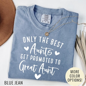 May include: A blue jean colored t-shirt with the text "ONLY THE BEST AUNTS GET PROMOTED TO Great Aunt" in white. A gold necklace with a pendant is draped across the shirt. The shirt is folded on a white surface.