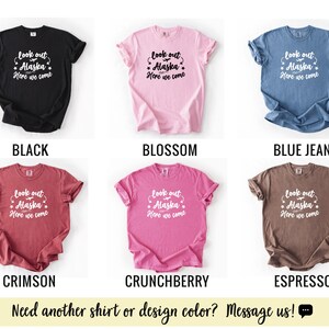 May include: A collection of six t-shirts in various colors: black, blossom, blue jean, crimson, crunchberry, and espresso. Each shirt displays the text "look out Alaska, Here we come" in white script. The bottom of the image has the text "Need another shirt or design color? Message us!"