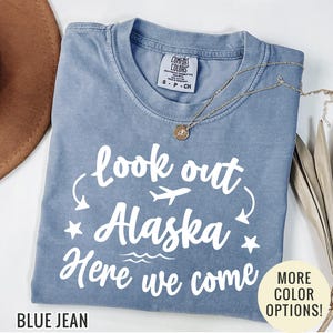 May include: A blue jean colored t-shirt with the text "look out Alaska Here we come" in white. The shirt has a crew neck and short sleeves. A gold necklace and a brown hat are also visible.