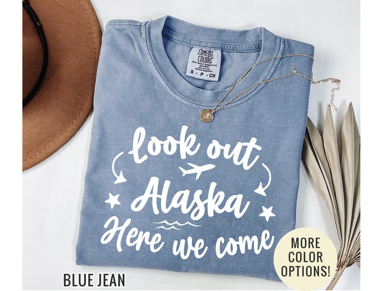 May include: A blue jean colored t-shirt with the text "look out Alaska Here we come" in white. The shirt has a crew neck and short sleeves. A gold necklace and a brown hat are also visible.
