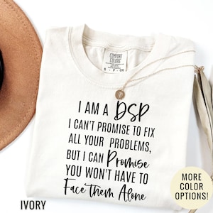May include: An ivory t-shirt with black text that reads: I AM A DSP I CAN'T PROMISE TO FIX ALL YOUR PROBLEMS, BUT I CAN PROMISE YOU WON'T HAVE TO FACE THEM ALONE. The shirt features a Comfort Colors brand label at the neck and a small circular pendant.