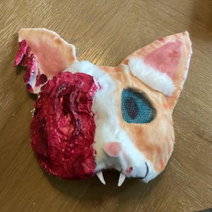 May include: A handcrafted cat mask featuring a white and orange fur-like texture, with one side covered in a textured, deep red material. The mask includes a single blue mesh eye opening, a small pink nose, and two white fangs protruding from the bottom.