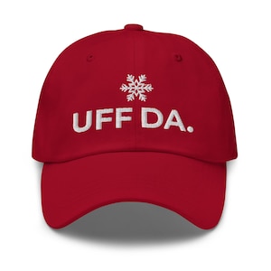 May include: A red baseball cap with the embroidered text "UFF DA." above a white snowflake design. The hat is a classic six-panel design with a curved brim and a fabric-covered adjustable closure.