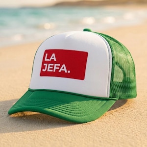 May include: A green and white trucker cap featuring a solid red rectangular patch on the front panel. The patch displays the white text LA JEFA. The cap has a green mesh back, a green brim with stitching, and a white foam front panel structure.
