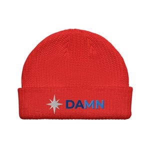 May include: A red knitted beanie hat with a turned-up brim. The hat has a white starburst emblem and the word "DAMN" embroidered in blue. The beanie is made of a soft, textured fabric.