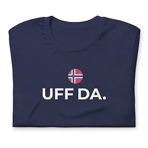 May include: Navy blue t-shirt with the text "UFF DA." in white, and a small Norwegian flag graphic. The shirt is folded and appears to be made of soft material.
