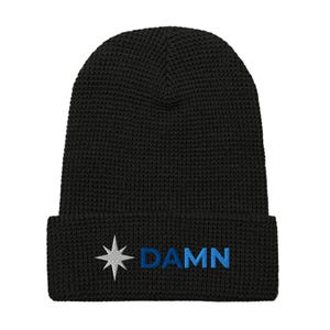May include: Black knitted beanie hat with a folded brim. The hat has a white and blue embroidered design of a star and the word "DAMN" in blue. The beanie is made of a textured knit material.