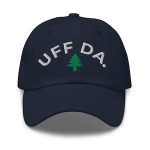 May include: A navy blue baseball cap with the embroidered text "UFF DA." in white, arched above a green pine tree graphic. The cap has a curved brim and a structured crown.