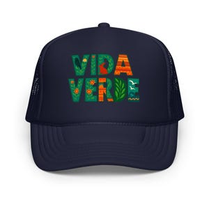 May include: Navy blue trucker hat featuring the words "VIDA VERDE" in decorative, colourful lettering. The letters are filled with floral and nature-themed illustrations, including a bird, rabbit, and flowers. The hat has a mesh back.