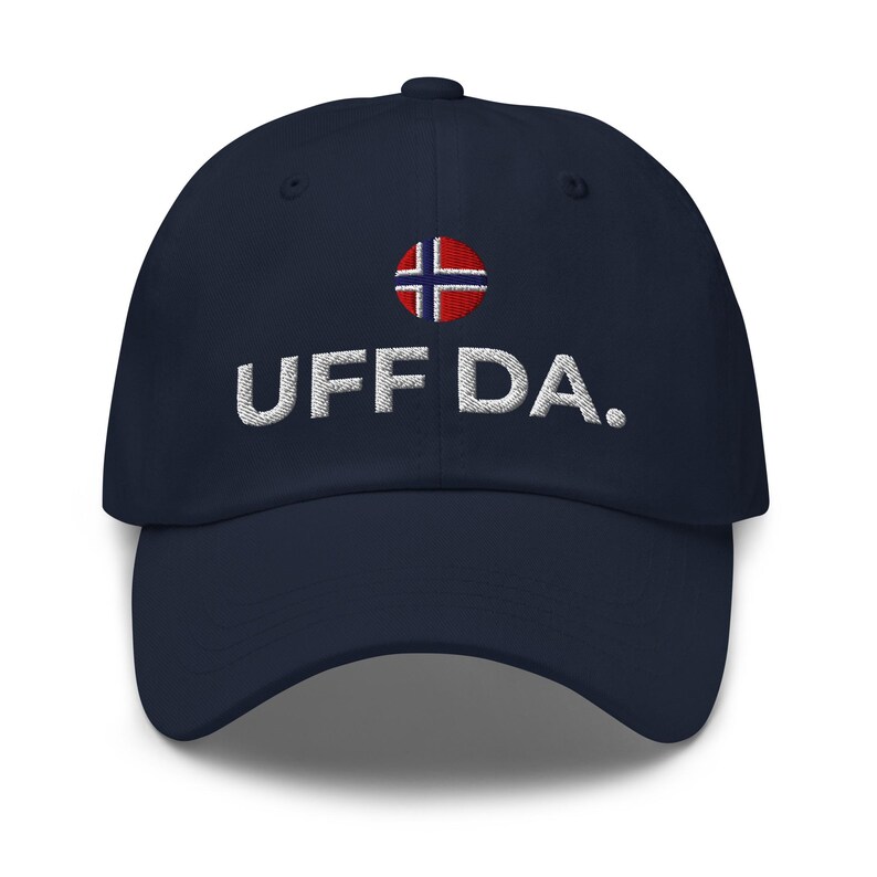 May include: Navy blue baseball cap with the embroidered text "UFF DA." above the Norwegian flag. The hat has a curved brim and a fabric strap closure on the back.