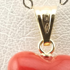 K&ouml;nnte beinhalten: Nahaufnahme eines goldenen Anh&auml;ngers mit einem herzf&ouml;rmigen roten Anh&auml;nger. Der Anh&auml;nger hat eine goldene &Ouml;se und eine kleine Schlaufe, die ihn mit dem Herzen verbindet. Die goldene &Ouml;se tr&auml;gt die Aufschriften "NA" und "750".