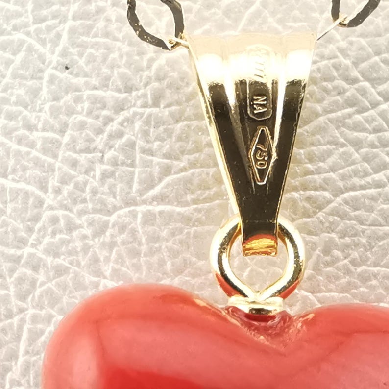 K&ouml;nnte beinhalten: Nahaufnahme eines goldenen Anh&auml;ngers mit einem herzf&ouml;rmigen roten Anh&auml;nger. Der Anh&auml;nger hat eine goldene &Ouml;se und eine kleine Schlaufe, die ihn mit dem Herzen verbindet. Die goldene &Ouml;se tr&auml;gt die Aufschriften "NA" und "750".