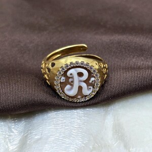 Op de afbeelding: Goudkleurige ring met een ovale camee met een witte letter "R". De cameo is omgeven door kleine, heldere edelstenen. De ringband heeft een decoratief, open ontwerp en is verstelbaar.