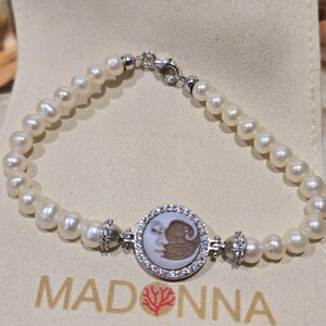 May include: A pearl bracelet with a silver clasp and a central circular charm. The charm features a carved design of a face within a circle of small, clear stones. The word "MADONNA" is printed below the bracelet.