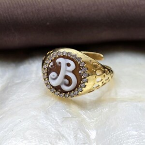Könnte beinhalten: Goldfarbener Ring mit braunem, ovalem Mittelteil mit einem weißen Buchstaben "B"-Design. Das Oval ist von kleinen, klaren Edelsteinen umgeben. Das Ringband hat ein strukturiertes Muster und liegt auf einer weißen Oberfläche.