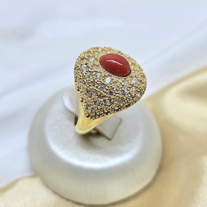 Könnte beinhalten: Ein goldener Ring mit einem ovalen, roten Korallenstein in der Mitte, umgeben von kleinen, klaren Edelsteinen. Der Ringkörper und die Fassung sind goldfarben und schaffen ein luxuriöses Design. Der Ring wird auf einem weißen Ständer präsentiert.