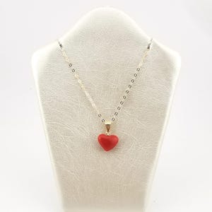 K&ouml;nnte beinhalten: Eine goldfarbene Halskette mit einer filigranen Kette und einem roten herzf&ouml;rmigen Anh&auml;nger. Die Halskette wird auf einem wei&szlig;en Schmuckst&auml;nder pr&auml;sentiert, der die leuchtende Farbe und das Design des Anh&auml;ngers hervorhebt.