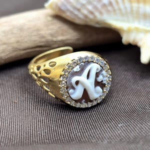 Pode incluir: Um anel dourado com um design de camafeu com a letra "A" em branco. O anel tem uma banda decorativa com detalhes recortados e é circundado por pequenas pedras preciosas transparentes. O camafeu está sobre um fundo castanho.