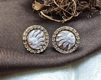 May include: A pair of round earrings featuring a carved sun design. The sun is white, set against a brown background, and surrounded by a ring of small, clear gemstones. The earrings have a gold-colored metal setting.