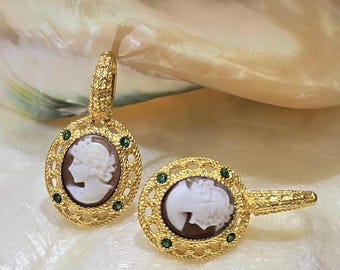 May include: A pair of gold-toned cameo earrings. Each earring features an oval cameo with a white profile against a brown background, surrounded by a gold filigree frame and accented with small green gemstones.
