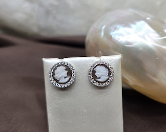 May include: A pair of stud earrings featuring a cameo design. Each earring has a brown oval cameo with a white profile, surrounded by a halo of small, clear crystals. The earrings are displayed on a white stand.
