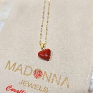 K&ouml;nnte beinhalten: Eine goldene Halskette mit einem herzf&ouml;rmigen Anh&auml;nger aus roter Koralle. Die Halskette wird auf einem beigen Beutel mit dem Text "MADONNA JEWELS" und einem Korallenast-Logo pr&auml;sentiert.