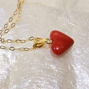 K&ouml;nnte beinhalten: Eine goldene Halskette mit einem herzf&ouml;rmigen roten Anh&auml;nger. Der Anh&auml;nger hat eine leuchtend rote Farbe und eine glatte, polierte Oberfl&auml;che. Die Kette ist zart und goldfarben, mit einem einfachen Kettendesign. Die Halskette wird auf einer wei&szlig;en, strukturierten Oberfl&auml;che pr&auml;sentiert.