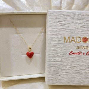 K&ouml;nnte beinhalten: Eine goldfarbene Halskette mit einem roten herzf&ouml;rmigen Anh&auml;nger, pr&auml;sentiert in einer offenen wei&szlig;en Schachtel. Die Schachtel hat goldenen Text "MADONNA JEWEL Coralli e C."