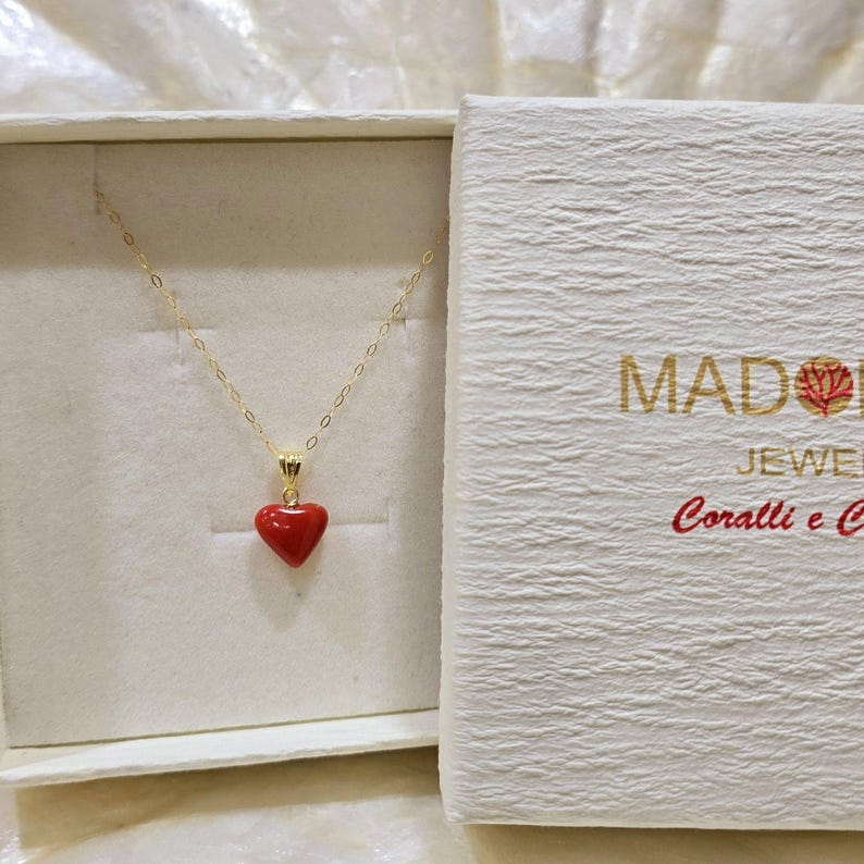 K&ouml;nnte beinhalten: Eine goldfarbene Halskette mit einem roten herzf&ouml;rmigen Anh&auml;nger, pr&auml;sentiert in einer offenen wei&szlig;en Schachtel. Die Schachtel hat goldenen Text "MADONNA JEWEL Coralli e C."